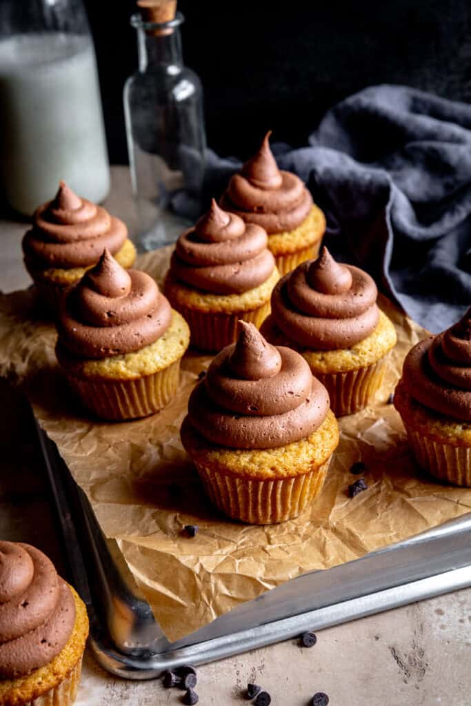 Vanilla cupcakes with chocolate frosting on a baking sheet and a jug of milk.