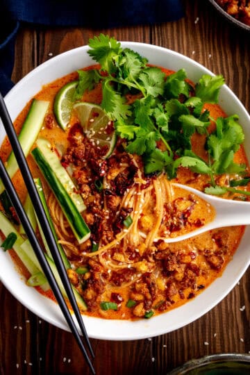 Bowl of vegan miso curry ramen topped with crispy tofu, cilantro, cucumber and lime wedges.