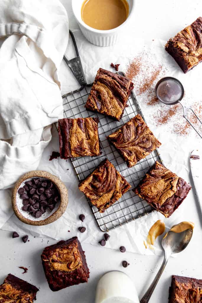 Peanut butter brownies on a wire rack, a cup of chocolate chips and a cup of peanut butter.