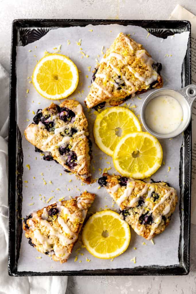 Lemon blueberry scones on a baking sheet with lemon slices and a cup of glaze.