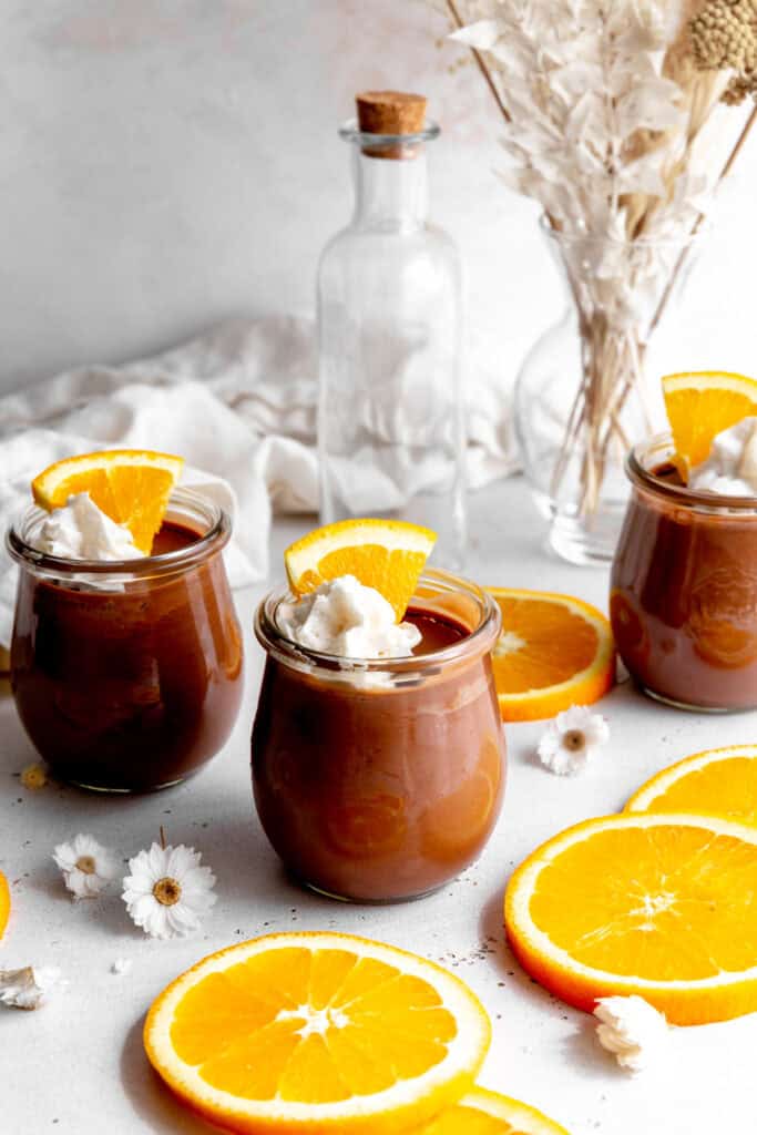 Glass jars of chocolate mousse, orange slices and small white flowers.