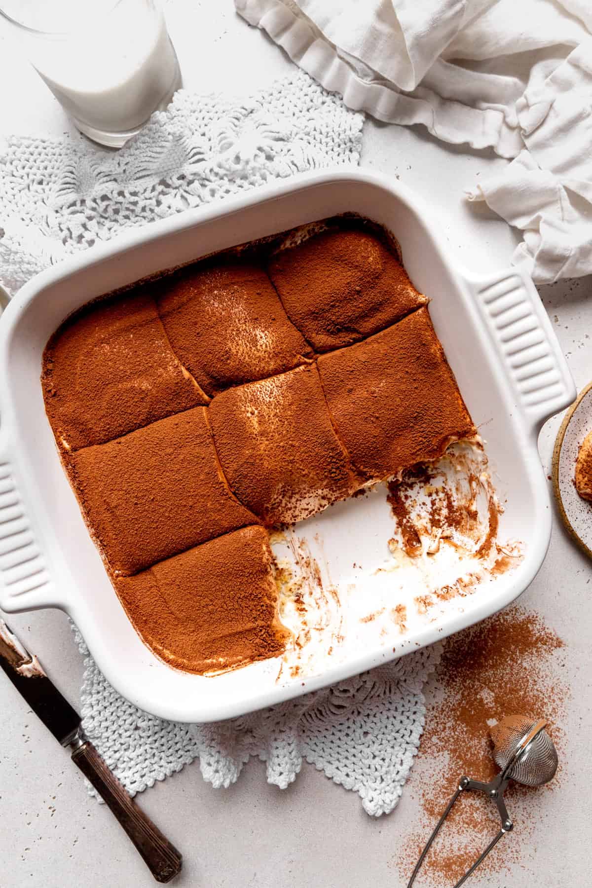 Tiramisu in a square dish, a glass of milk and a linen napkin.
