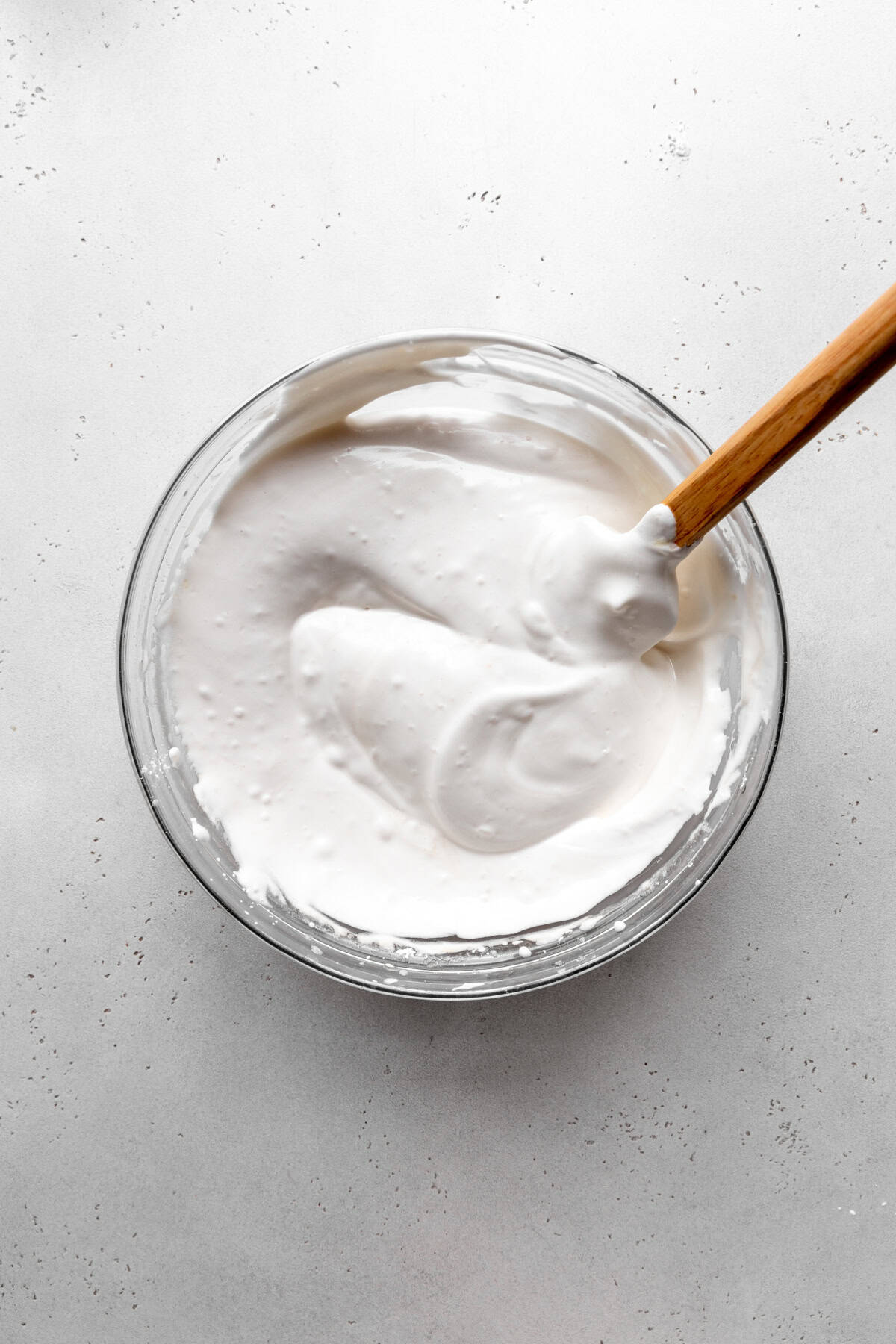 Glass bowl of Glass bowl of whipped aquafaba meringue and a rubber spatula.