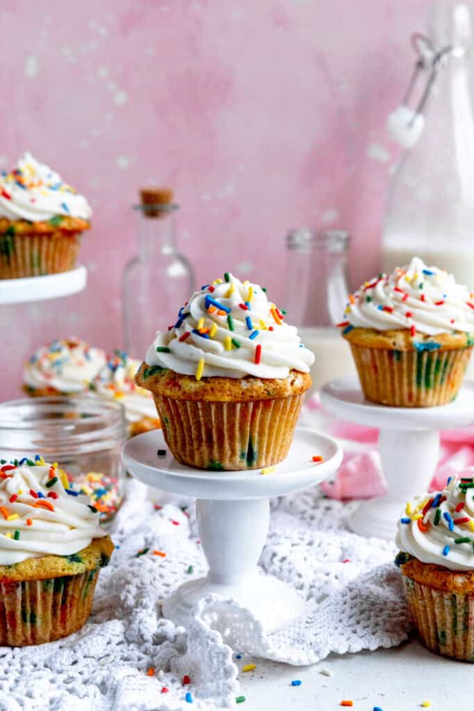 Funfetti cupcakes on white pedestals and a jug of milk.