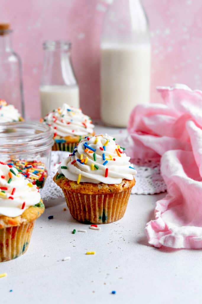 Funfetti cupcakes, a jar of sprinkles, a pink linen and a jug of milk.