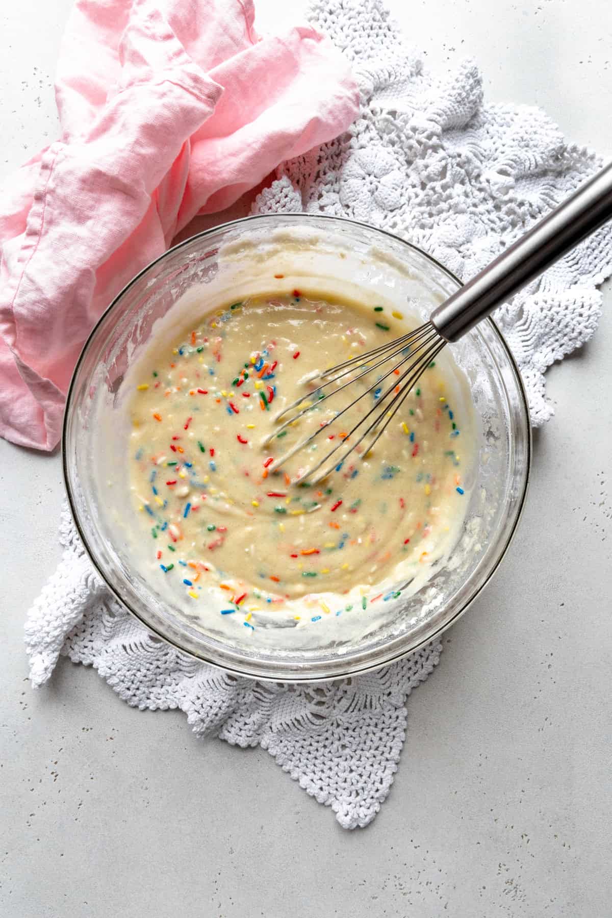 Funfetti cake batter in a glass bowl with a whisk and a pink napkin.