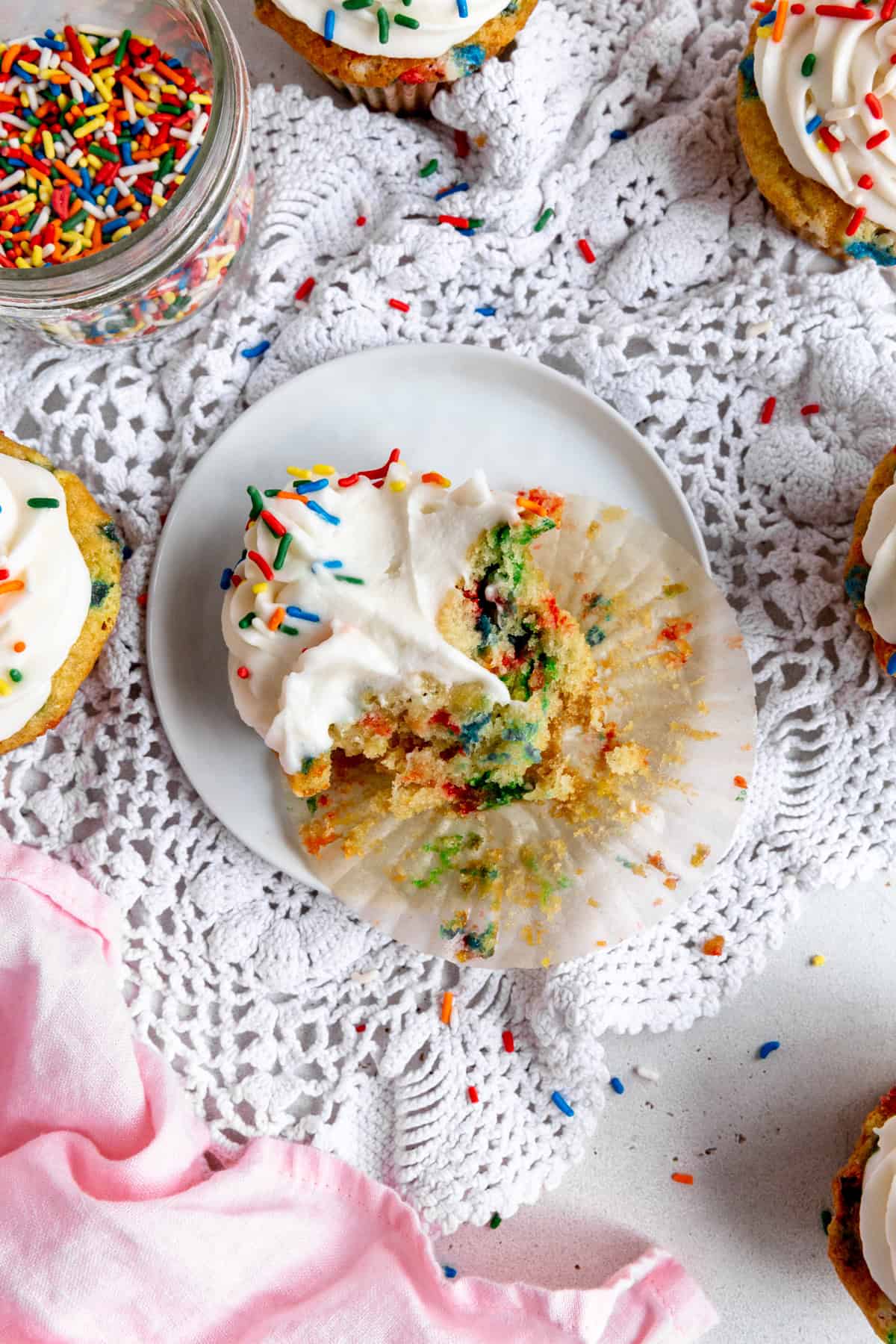 Partially eaten funfetti cupcake on a white plate and a jar of sprinkles.