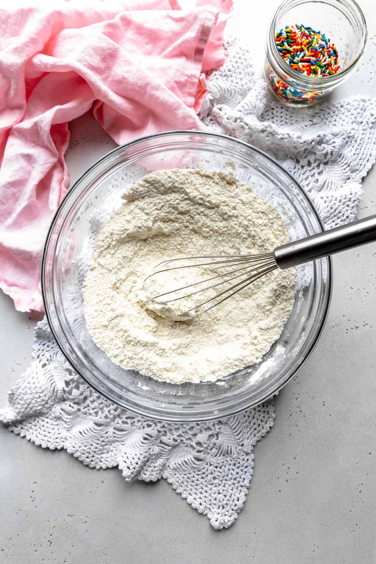 Glass bowl of flour with a whisk and a jar of multi-colored sprinkles.