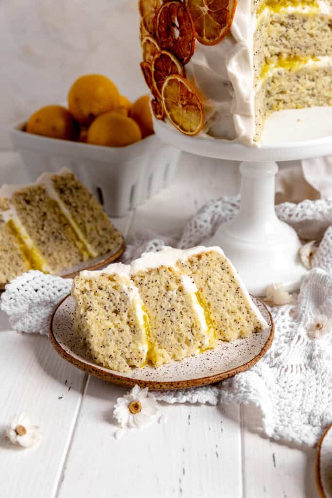 Slice of lemon poppyseed layer cake on a plate and a basket of lemons.