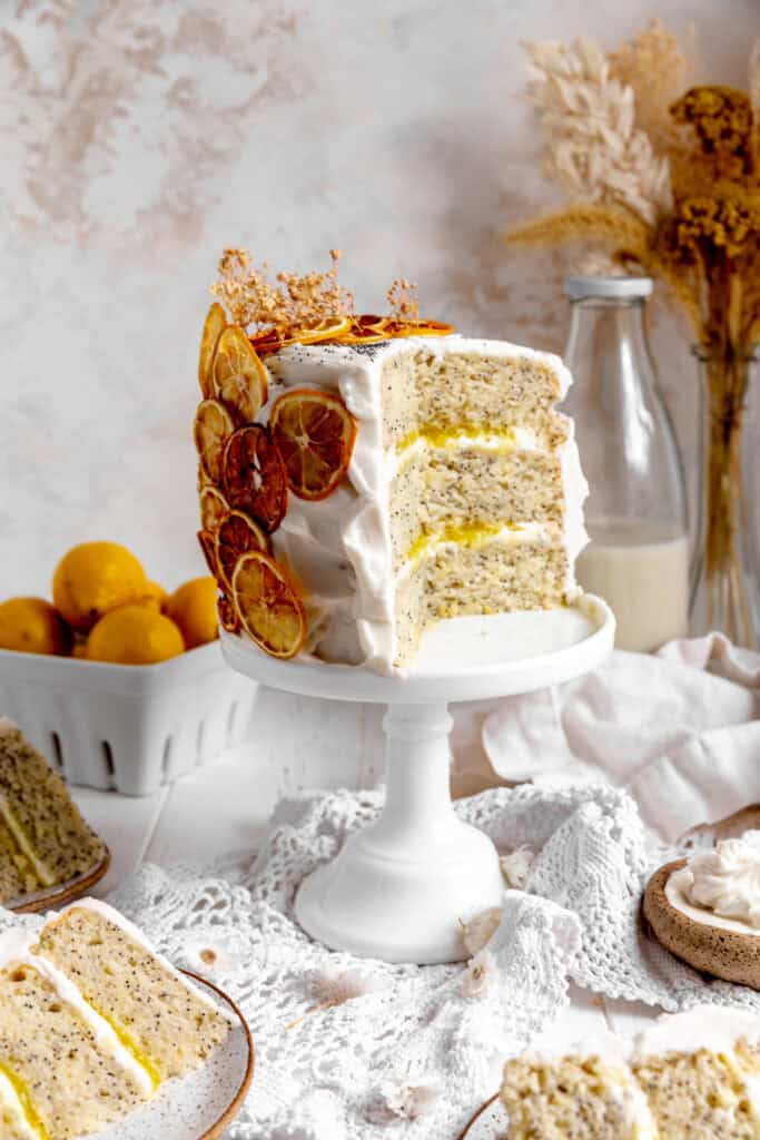 Lemon poppyseed layer cake on a cake stand, a basket of lemons and a jug of milk.
