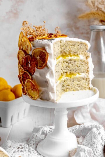 Lemon poppyseed layer cake on a cake stand, a basket of lemons and a jug of milk.