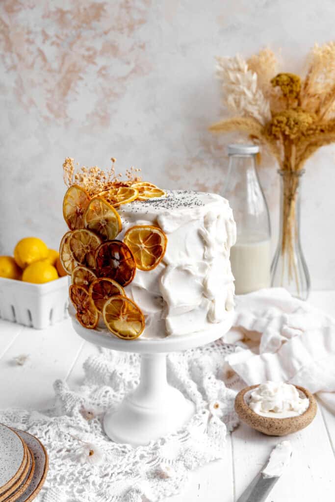 Lemon poppyseed cake on a cake stand, a stack of plates and a basket of lemons.