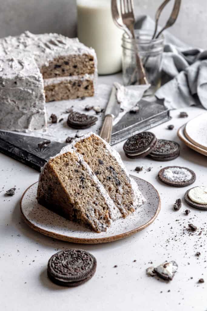 Slice of oreo cake on a plate, a knife covered in frosting and a jug of milk.