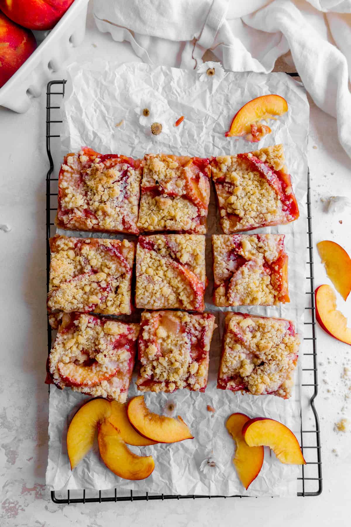 Peach crumble bars on a wire rack, a linen napkin and a basket of peaches.