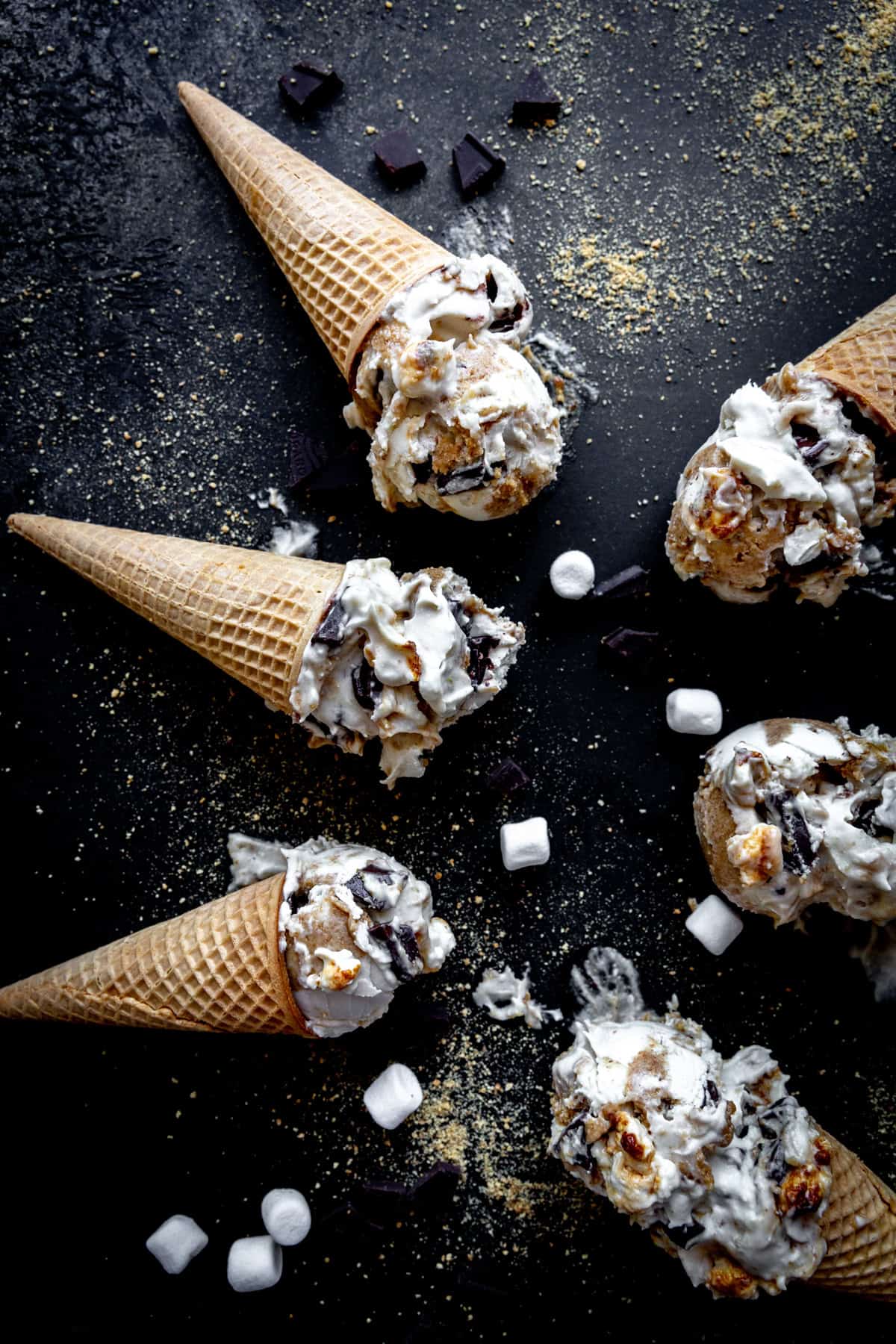 S'mores ice cream cones laying on a black surface with marshmallows and chocolate chunks.