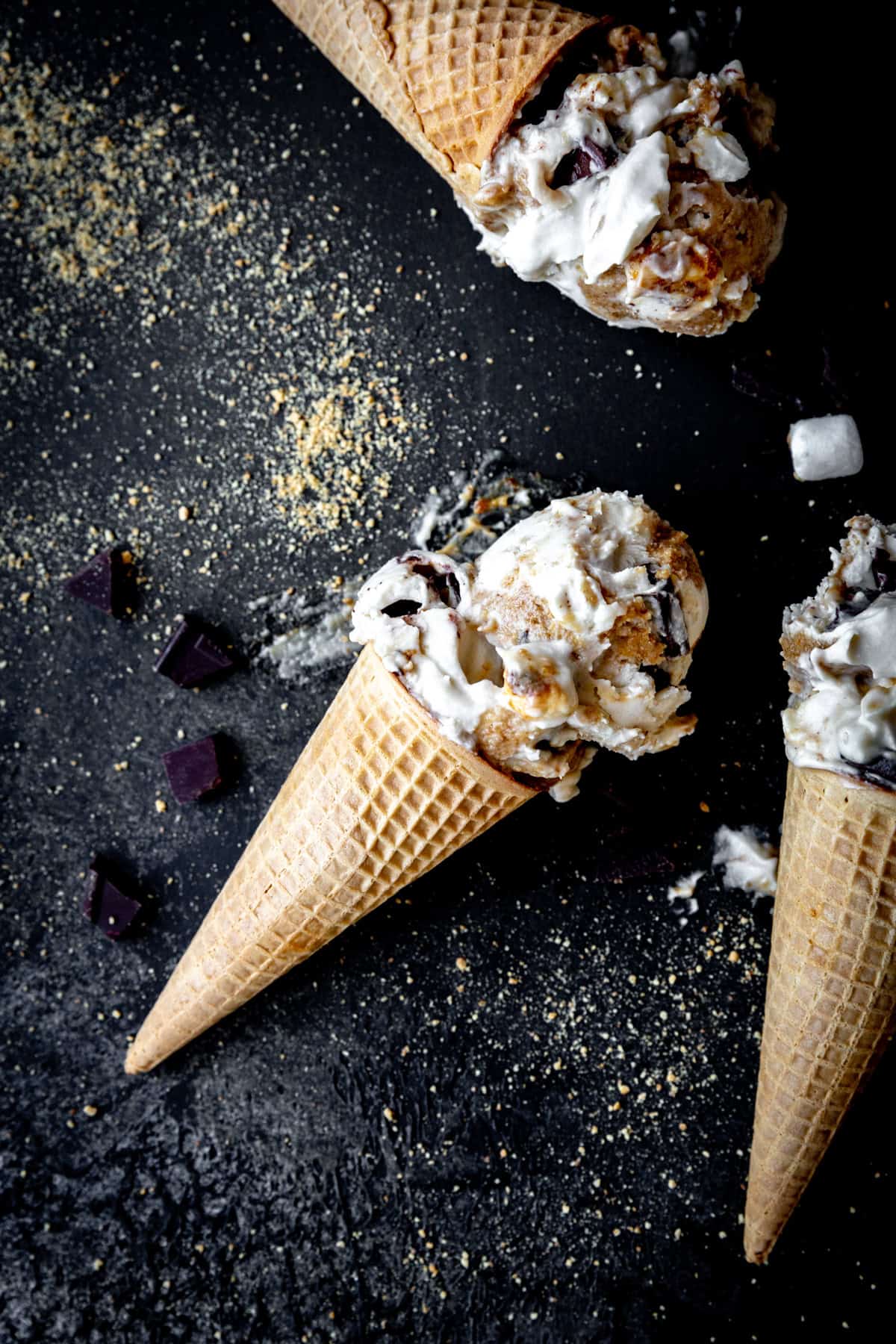 S'mores ice cream cones laying on a black background covered in graham cracker crumbs.