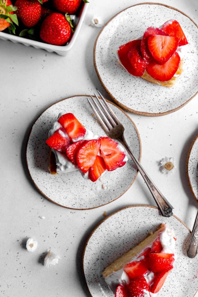 Slices of strawberry shortcake cake on white plates with forks and a basket of strawberries.