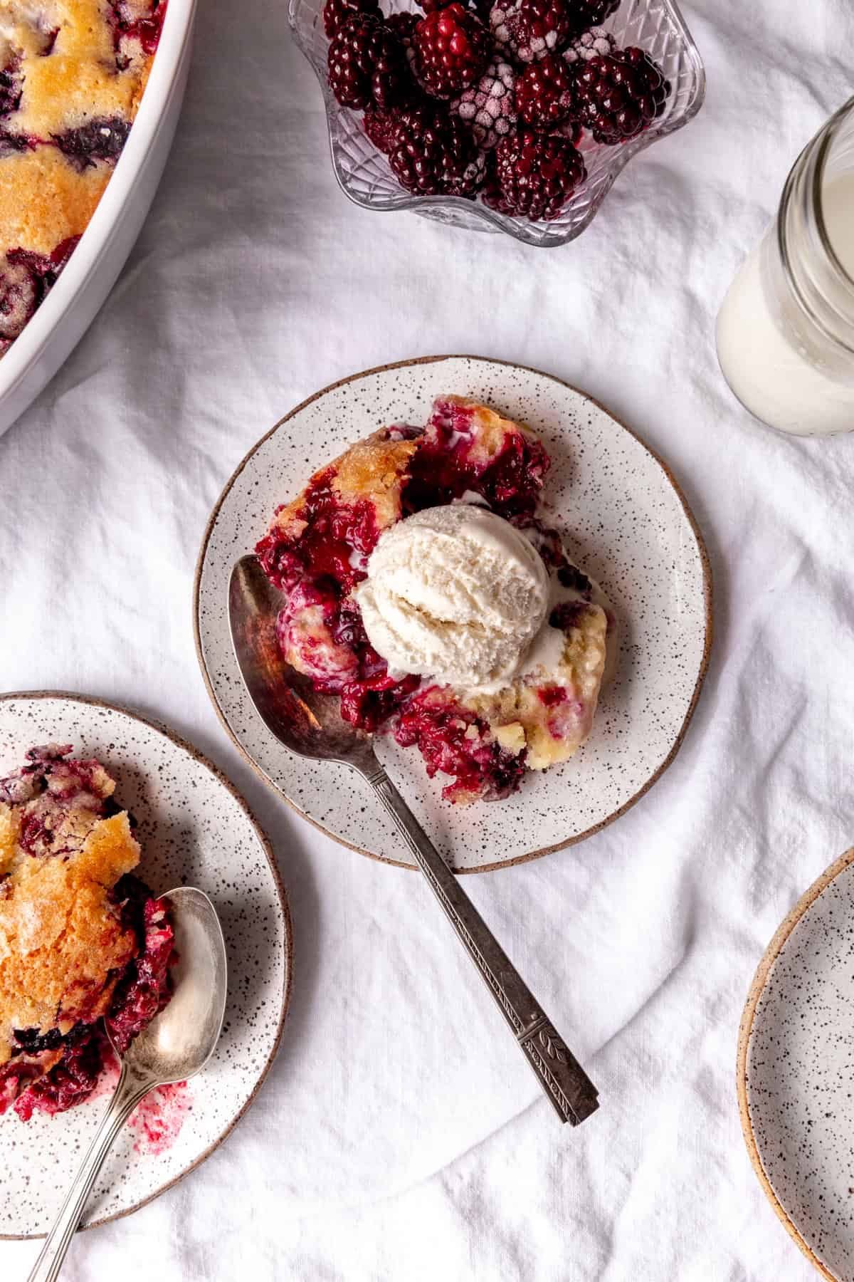 Plates of blackberry cobbler topped with ice cream, a bowl of blackberries and a glass of milk.