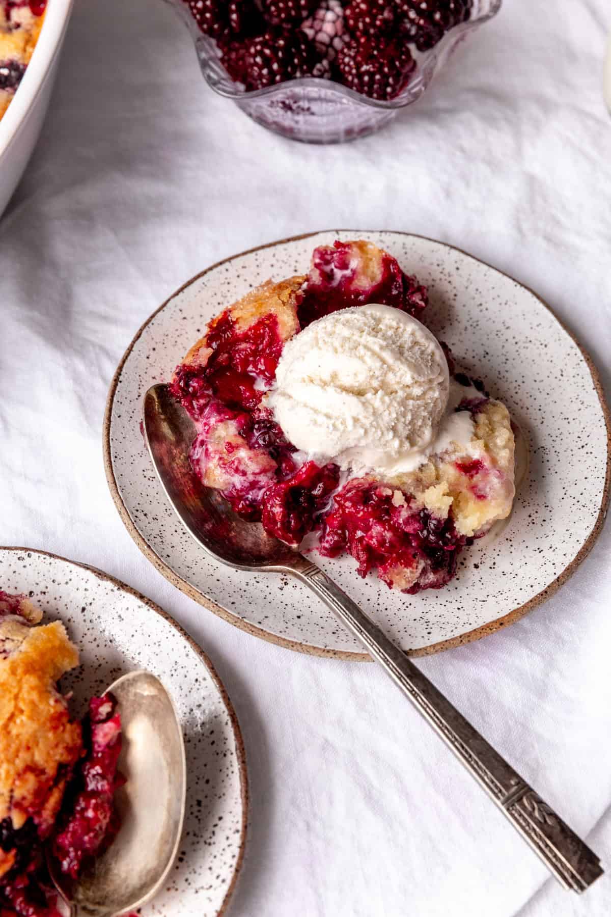 Spoon digging into a plate of blackberry cobbler on a white plate.