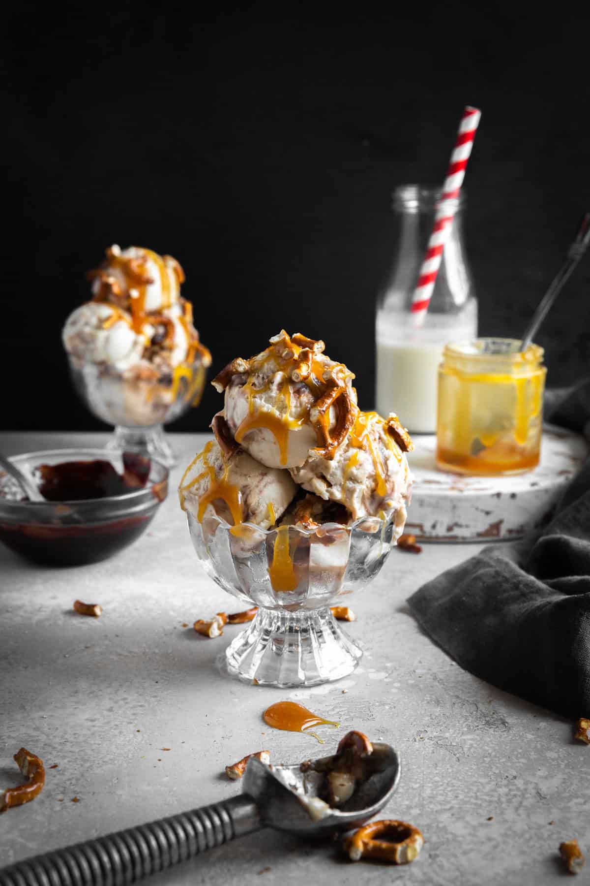 Two dishes of caramel pretzel ice cream, a bowl of fudge sauce and a jug of milk with a straw.