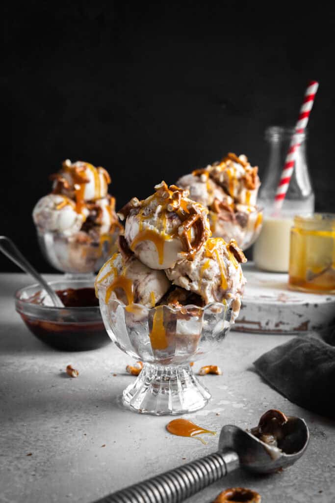 Glass dish of caramel pretzel ice cream, bowl of fudge sauce and an ice cream scoop.