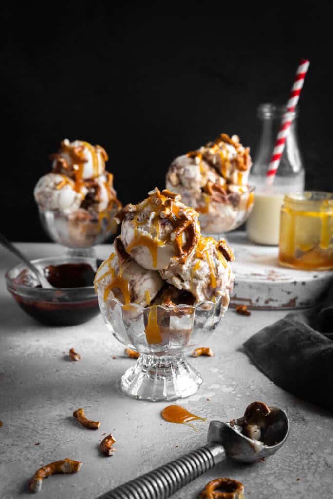 Three glass dishes of caramel pretzel ice cream, a jar of caramel sauce and a jug of milk.