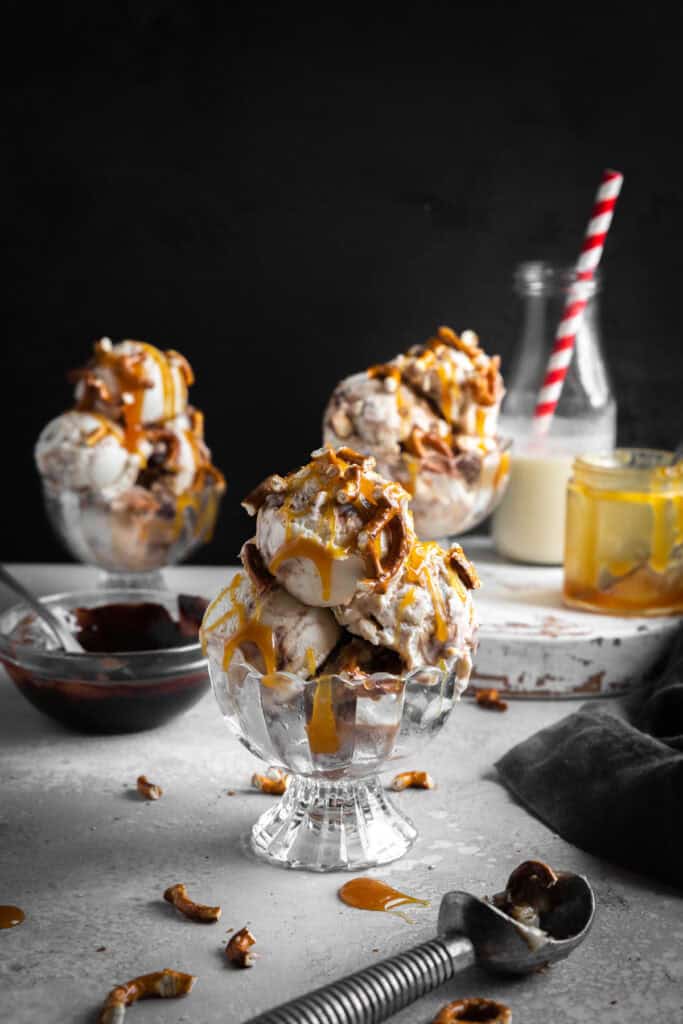 Three glass dishes of caramel pretzel ice cream, a jar of caramel sauce and a jug of milk.