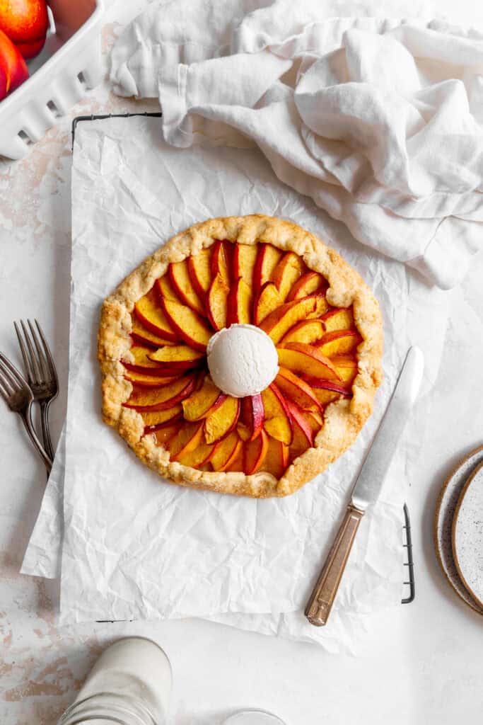 Nectarine galette on a baking sheet, a basket of nectarines, a glass of milk and a linen napkin.