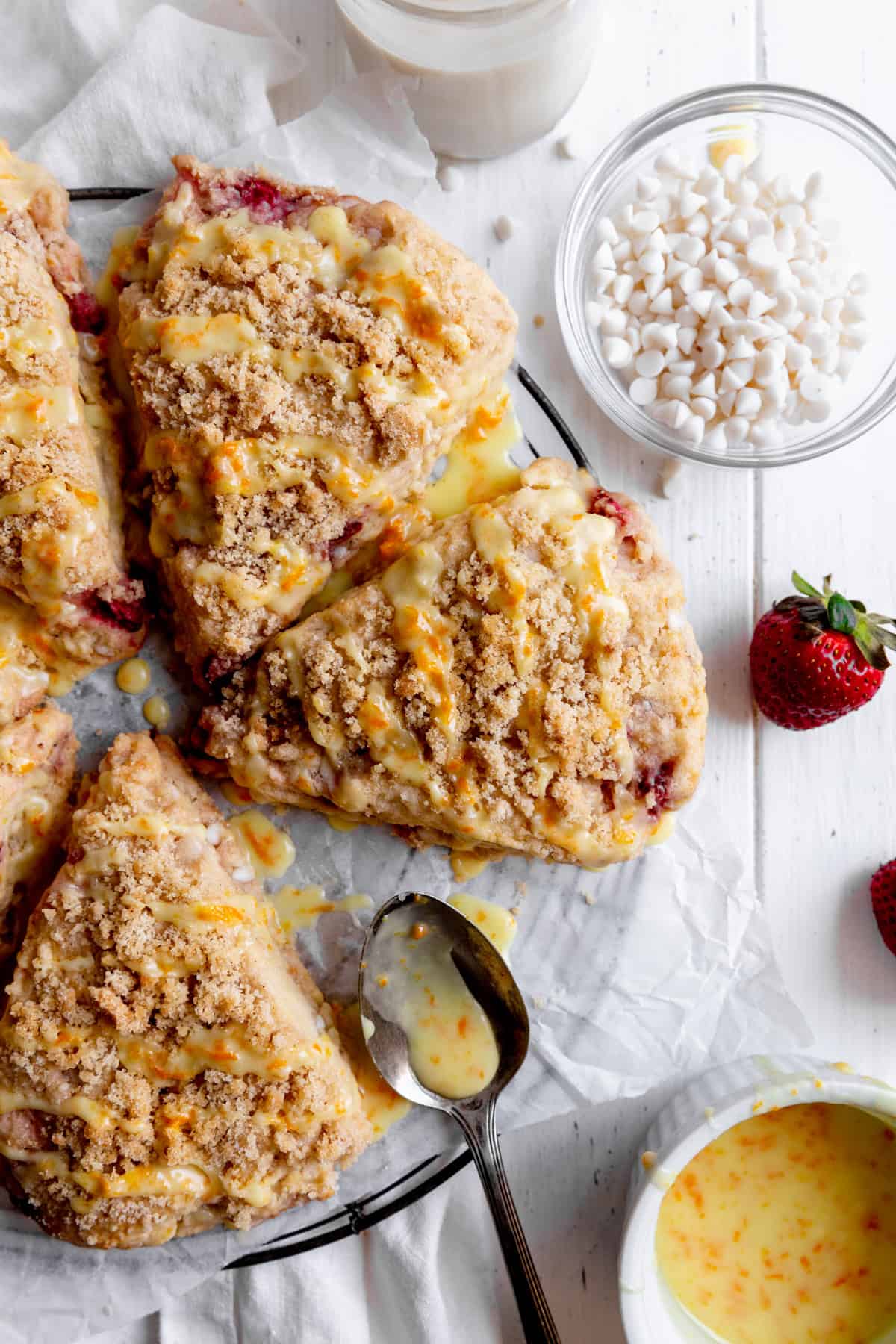 Strawberry rhubarb scones on a wire rack, a bowl of white chocolate chips and a bowl of orange glaze.