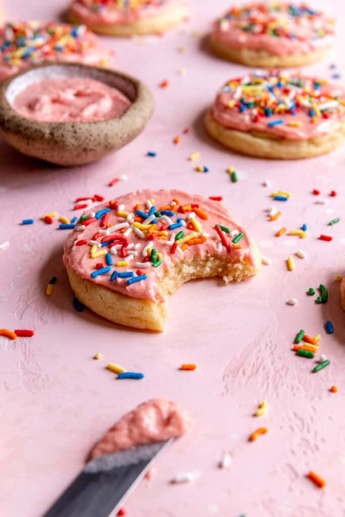 Sugar cookies on a pink surface with a knife and a small bowl of frosting.
