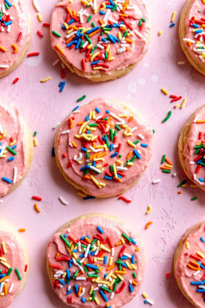 Sugar cookies on a pink surface topped with strawberry frosting and sprinkles.