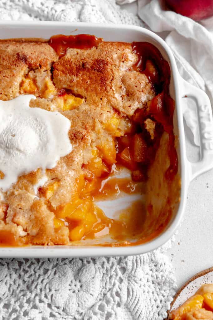 Half eaten peach cobbler covered in melted ice cream in a white baking dish.