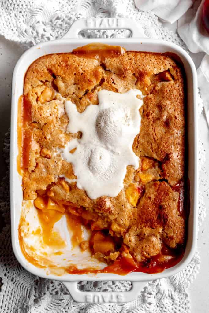 Half eaten peach cobbler covered in melted ice cream in a white baking dish.