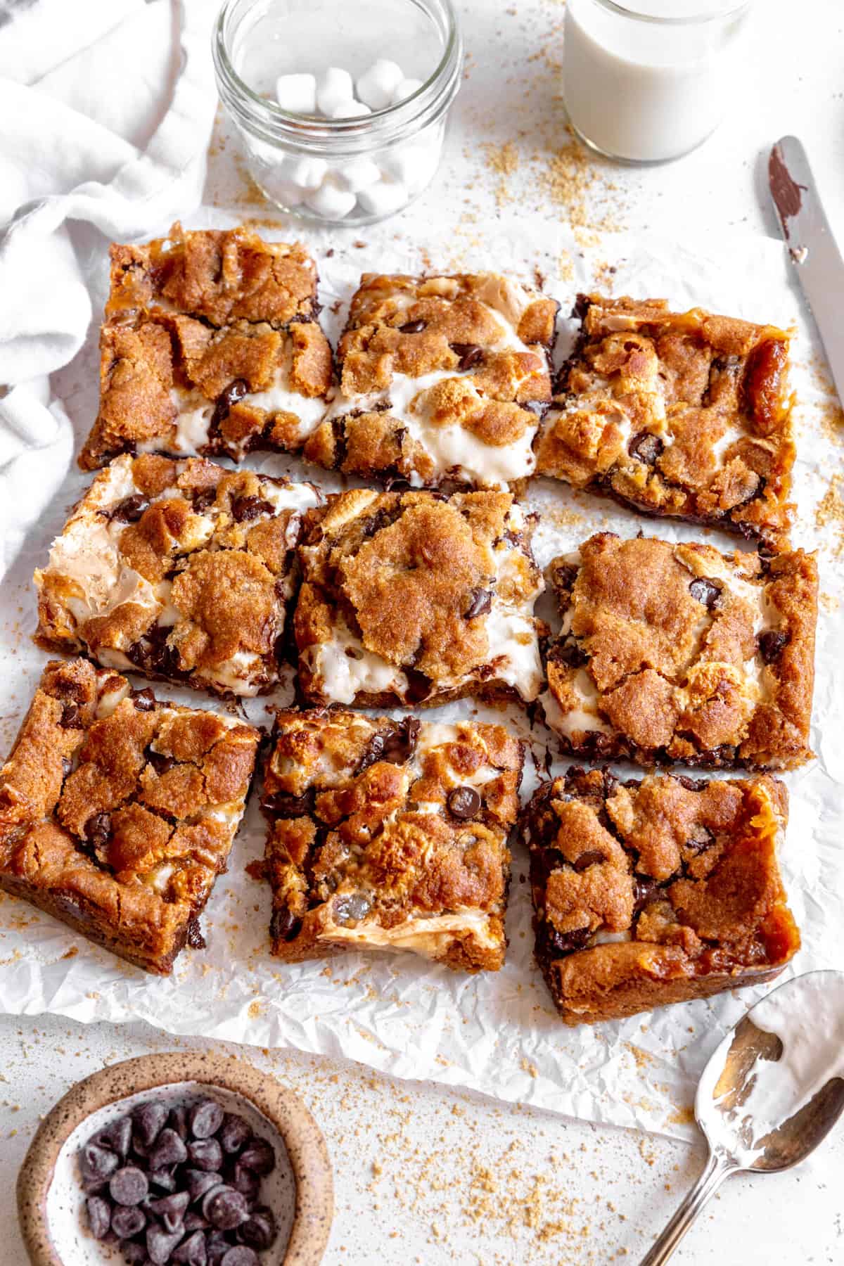 S'mores bars, a cup of chocolate chips, a cup of marshmallows and a glass of milk.