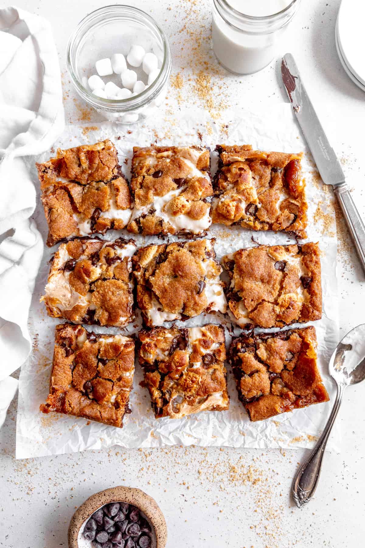 S'mores bars on a white surface with a cup of marshmallows and a cup of milk.