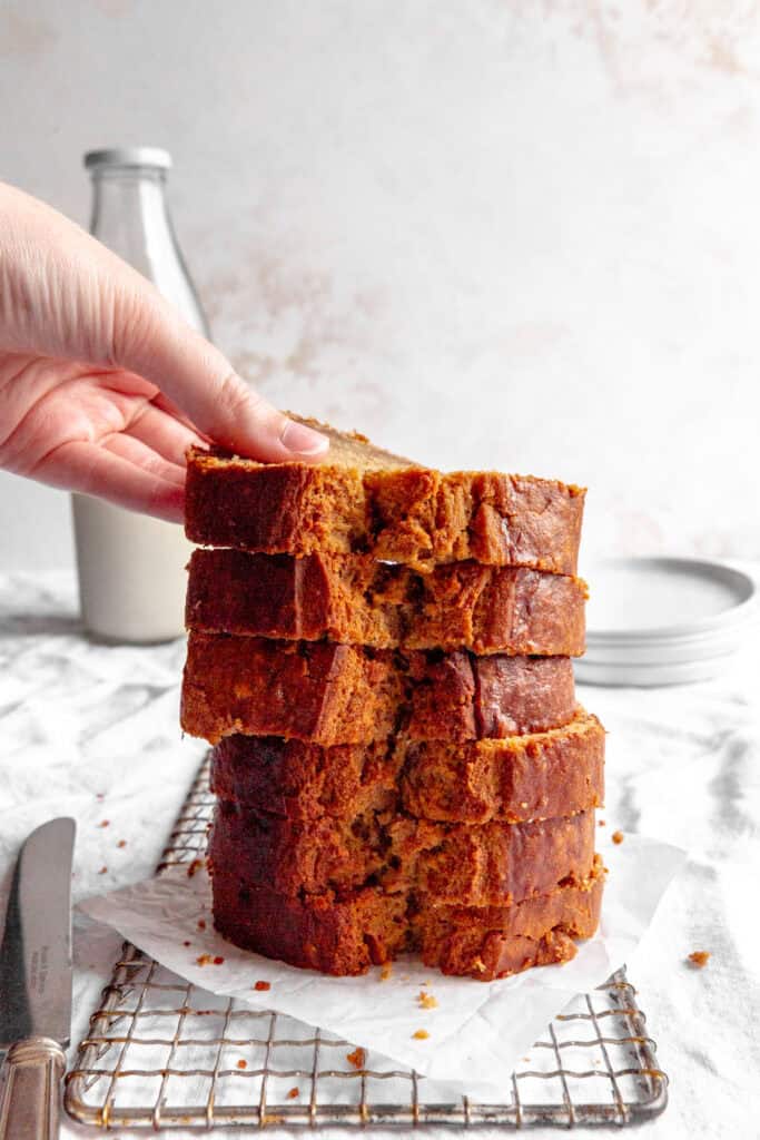 Hand lifting a slice of banana bread off of a stack and a jug of milk.