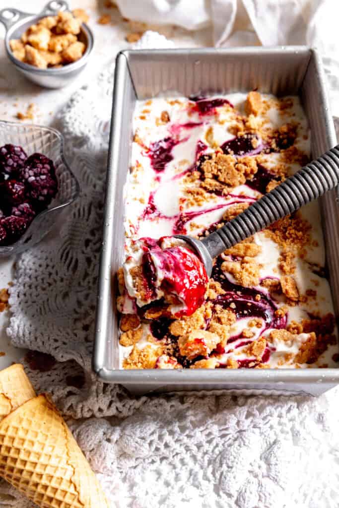 Ice cream scoop in a pan of blackberry cobbler ice cream and a cup of blackberries.