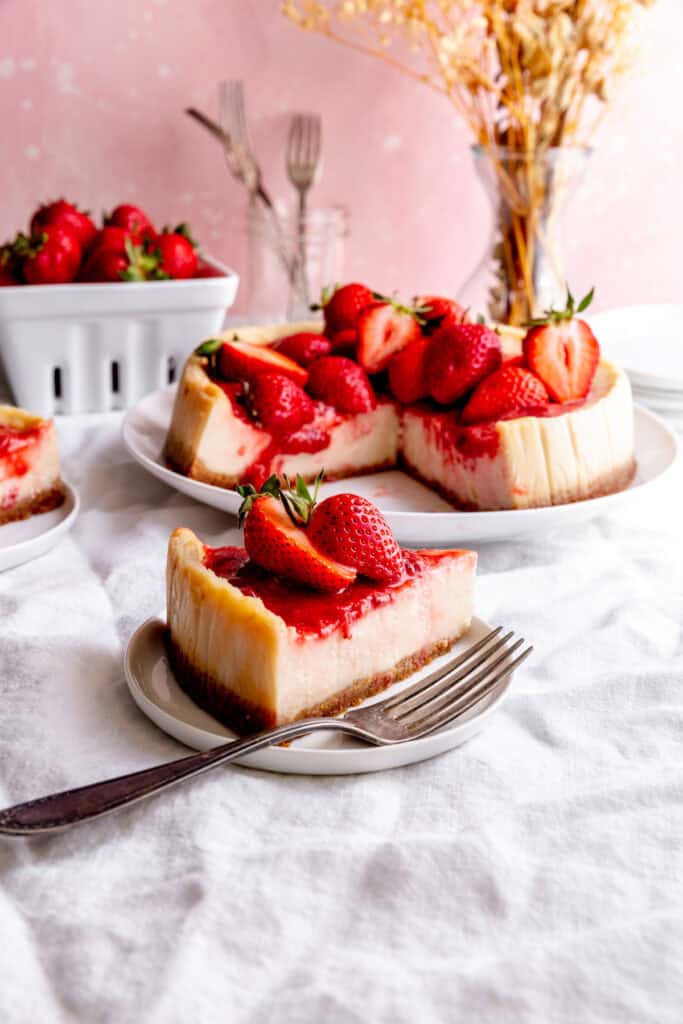 Slice of cheesecake on a plate with a fork and a basket of strawberries.