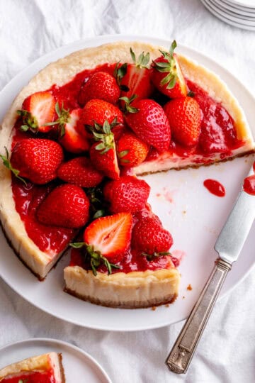 Cheesecake on a plate with slices missing and a knife covered in strawberry sauce.