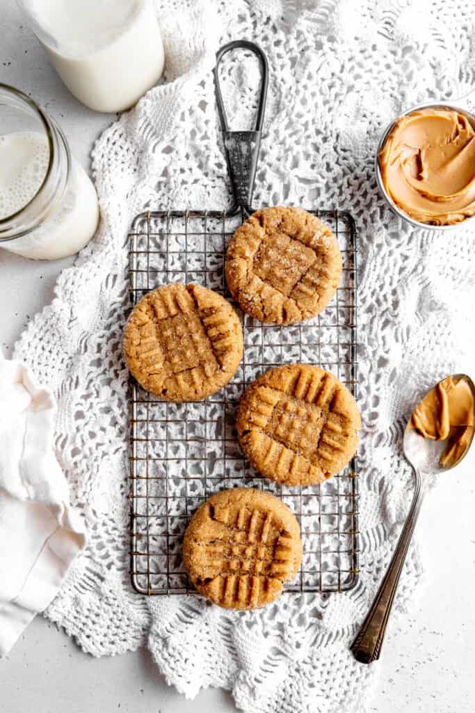 Peanut butter cookies on a wire rack, a cup of peanut butter and a glass of milk.