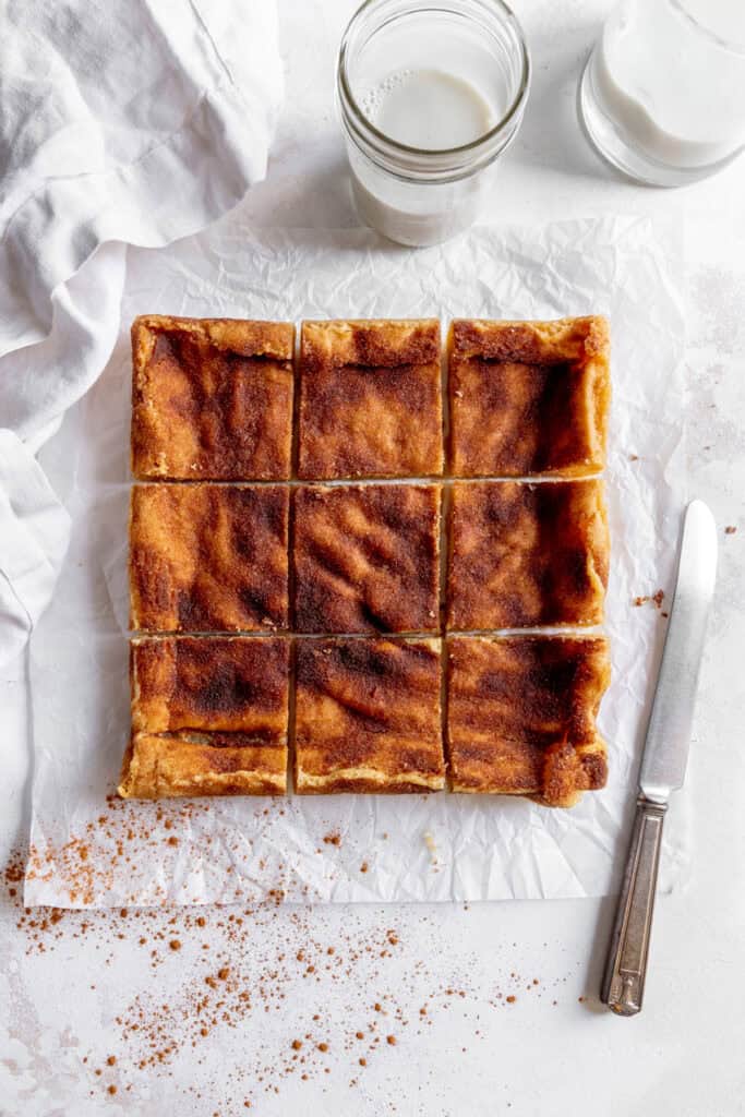 Sliced snickerdoodle bars on a white surface with a linen napkin and cups of milk.