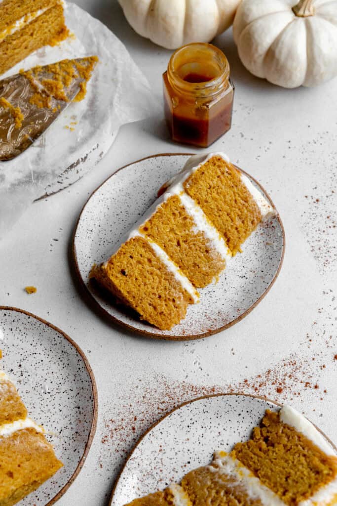 Slices of pumpkin cake on white plates, white pumpkins and a jar of caramel sauce.