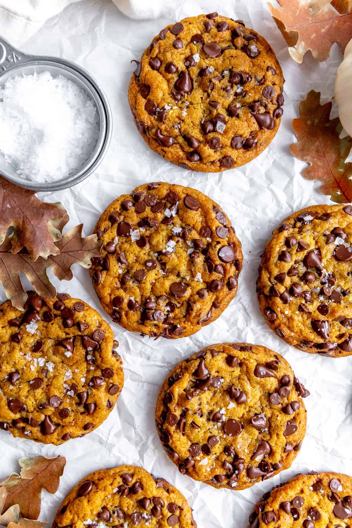 Pumpkin chocolate chip cookies on white parchment with a cup of sea salt.