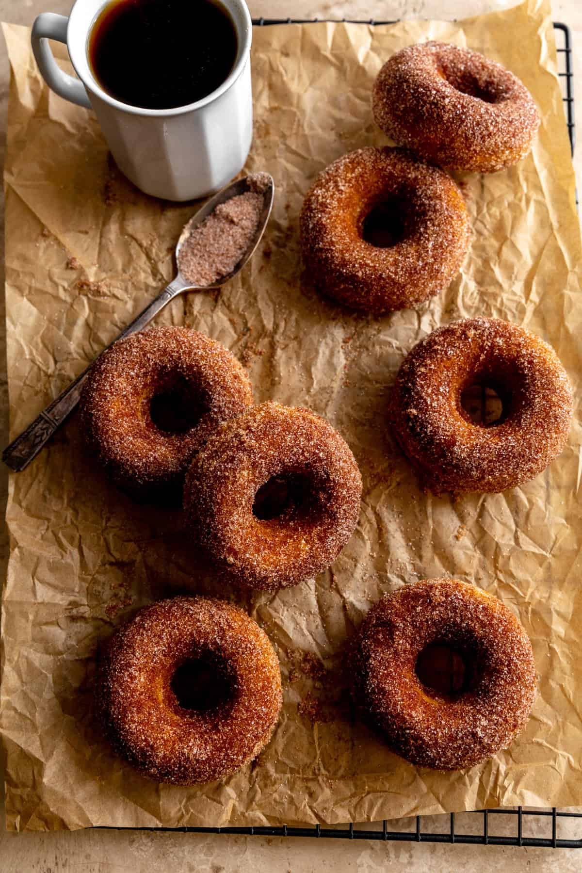 Pumpkin donuts on a cooling rack with a cup of coffee and a spoon of cinnamon sugar.